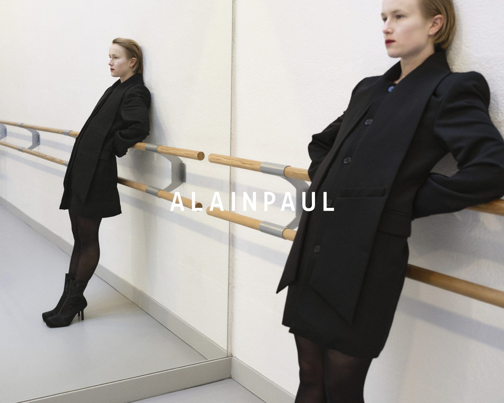 Model in black blazer and tights leaning against a ballet barre with mirror reflection for Alainpaul Fall 2024 campaign