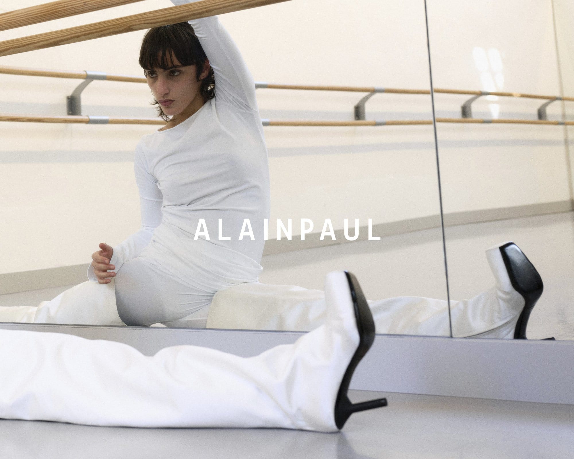 Model in white long-sleeve top and white trousers with heels stretches near a ballet barre and studio mirror in Alainpaul campaign.