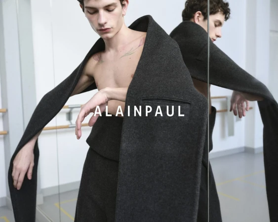 A shirtless male model wearing dark grey trousers draped with an oversized coat, standing before studio mirrors.