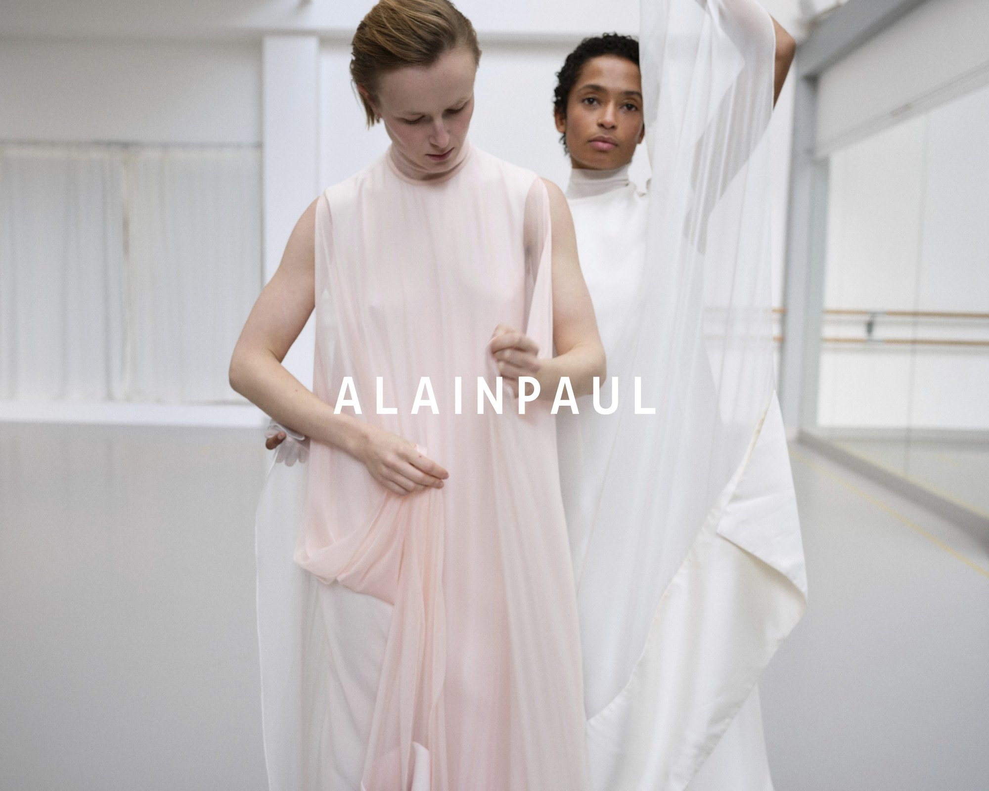 Two models in flowing white and pale pink garments posing in a bright minimalist studio space with the brand name Alainpaul overlaid.