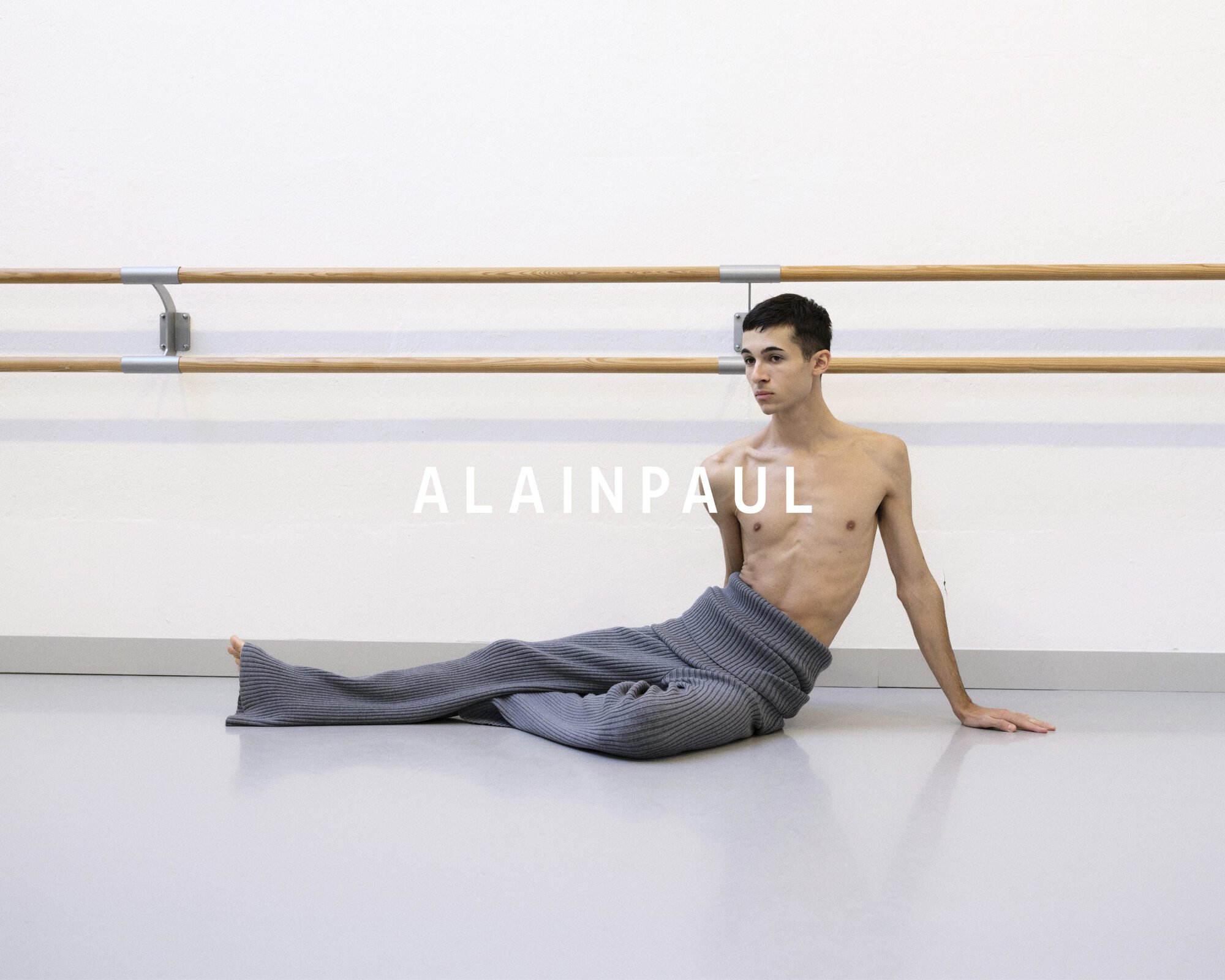 A shirtless male model wearing grey knit trousers sitting on a light studio floor in front of a wooden ballet barre