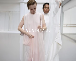 Two models in flowing white and pale pink garments posing in a bright minimalist studio space with the brand name Alainpaul overlaid.