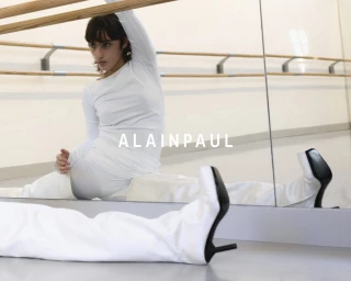 Model in white long-sleeve top and white trousers with heels stretches near a ballet barre and studio mirror in Alainpaul campaign.
