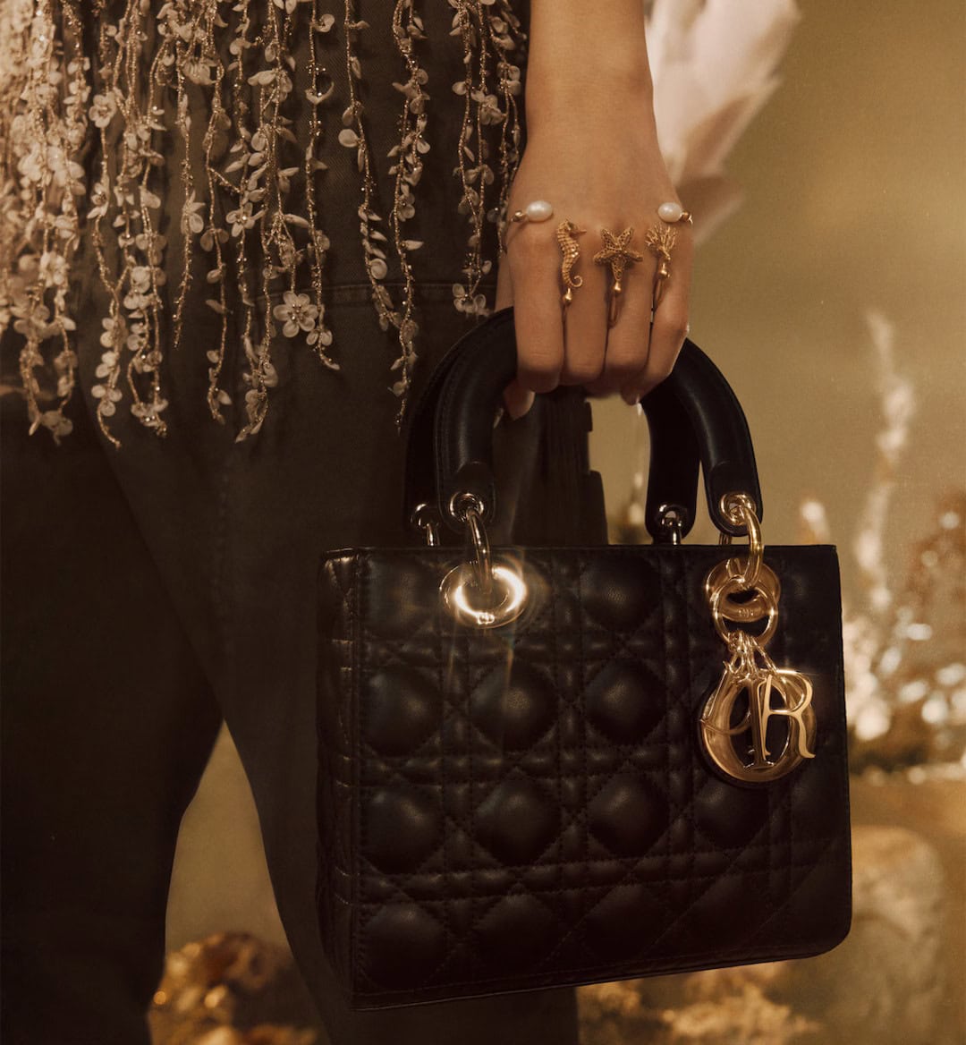 Close-up of a person holding a black quilted handbag with gold charms and wearing gold rings and a beaded top