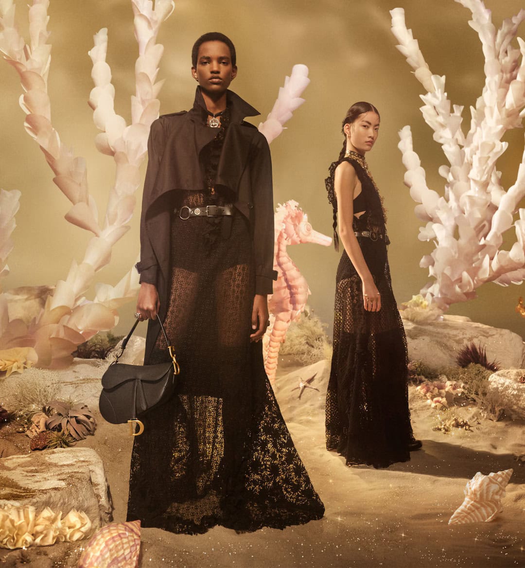 Two models in black lace dresses posing in a surreal underwater fantasy set with white coral and sandy ground for Dior