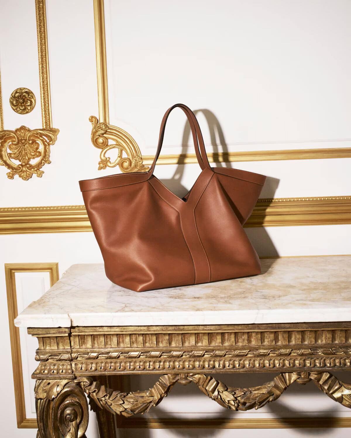 Brown leather tote bag resting on an ornate carved gilt table against a white wall with gold neoclassical moldings