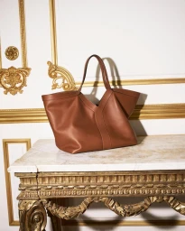 Brown leather tote bag resting on an ornate carved gilt table against a white wall with gold neoclassical moldings