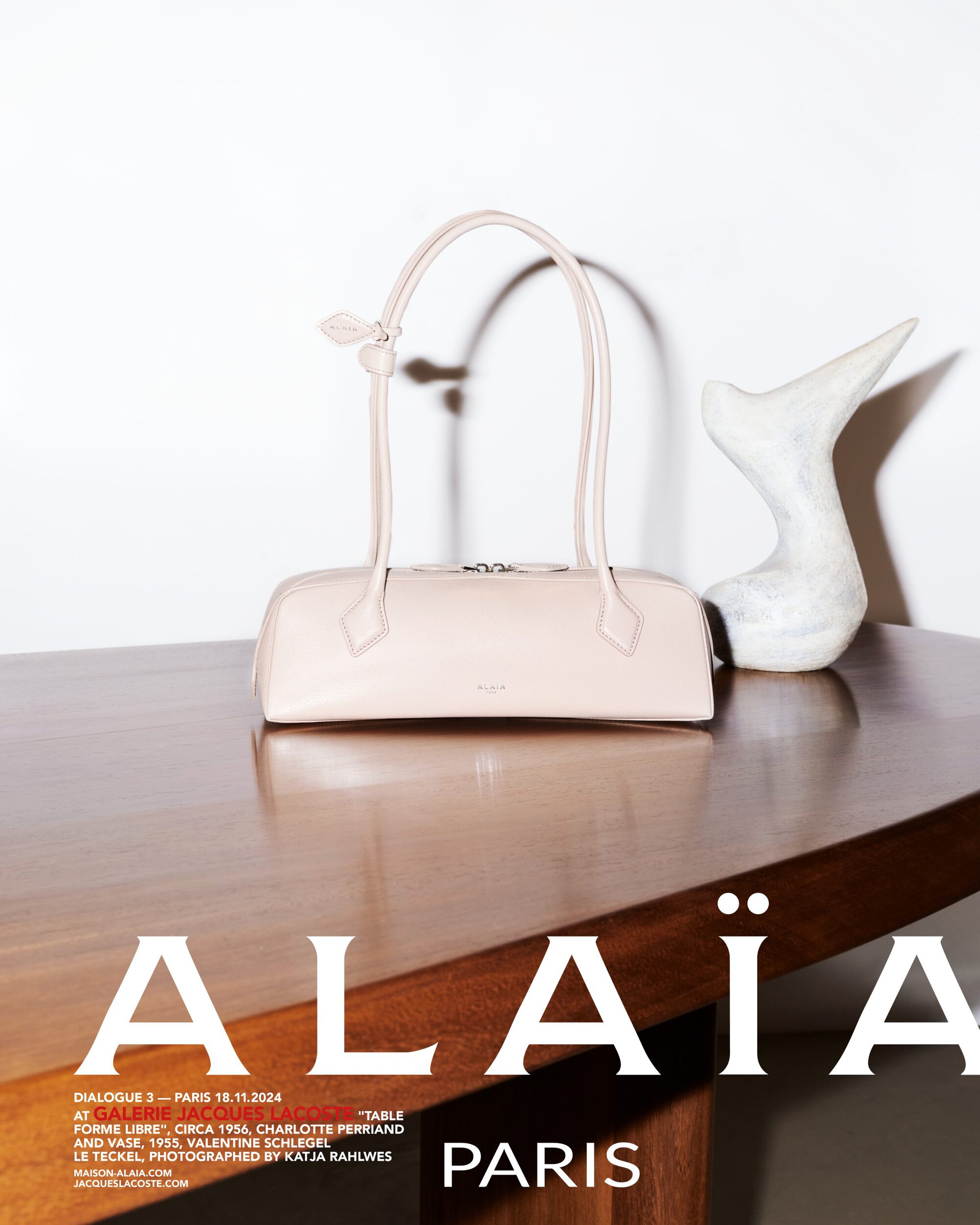A pale pink leather handbag sits on a polished wooden table next to a white sculptural art piece against a plain white wall.
