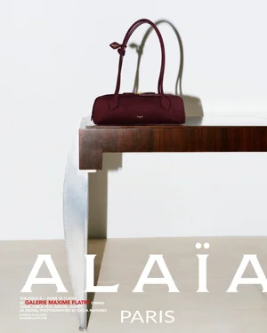 Burgundy leather handbag with long dual straps resting on a dark wood and metallic console table against a white wall.