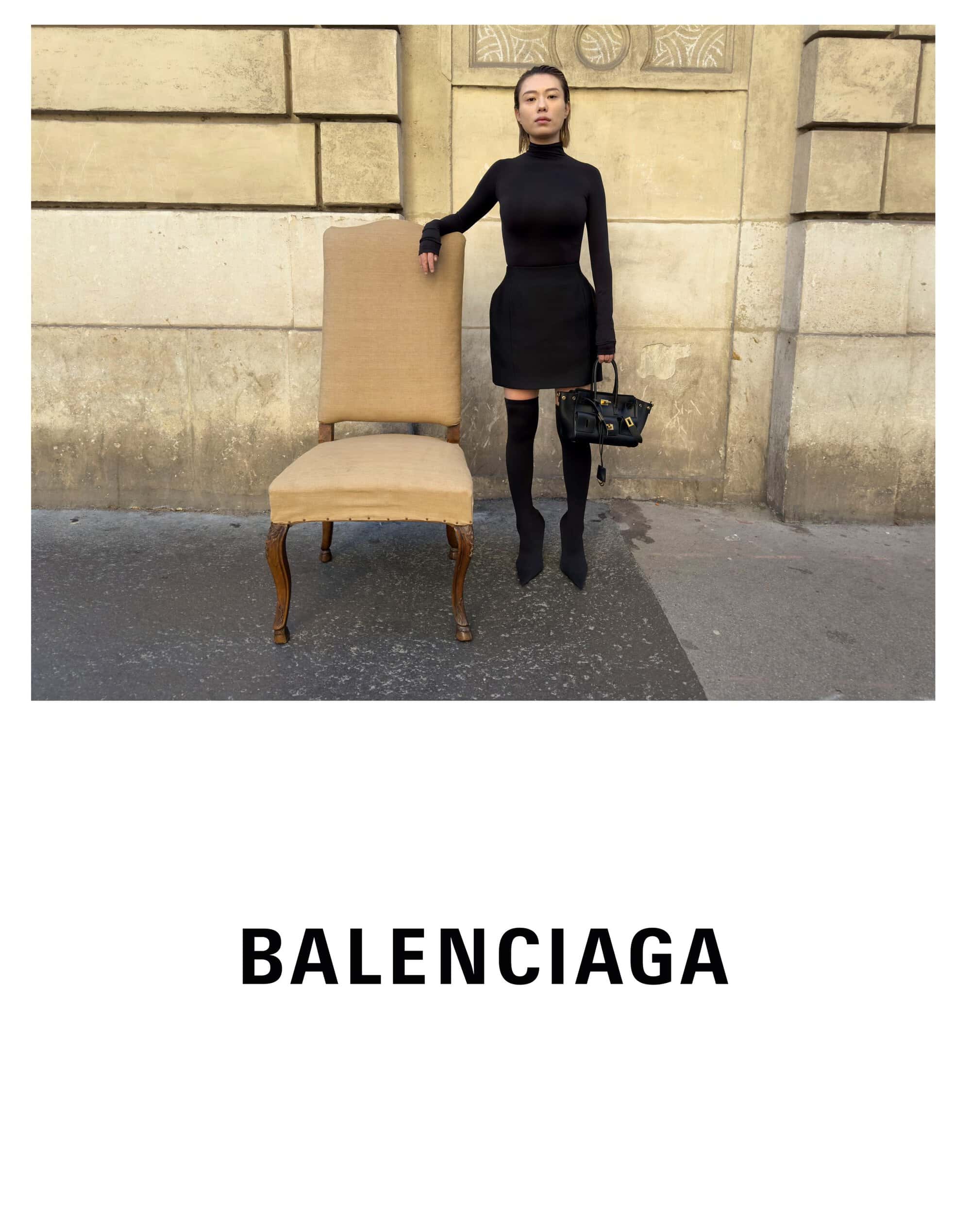 Model standing next to a large upholstered chair wearing all-black attire and holding a dark handbag outdoors in Paris