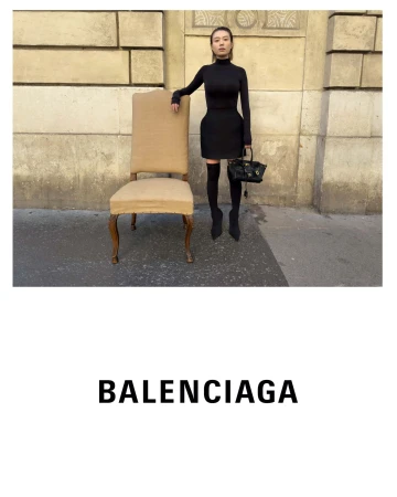 Model standing next to a large upholstered chair wearing all-black attire and holding a dark handbag outdoors in Paris