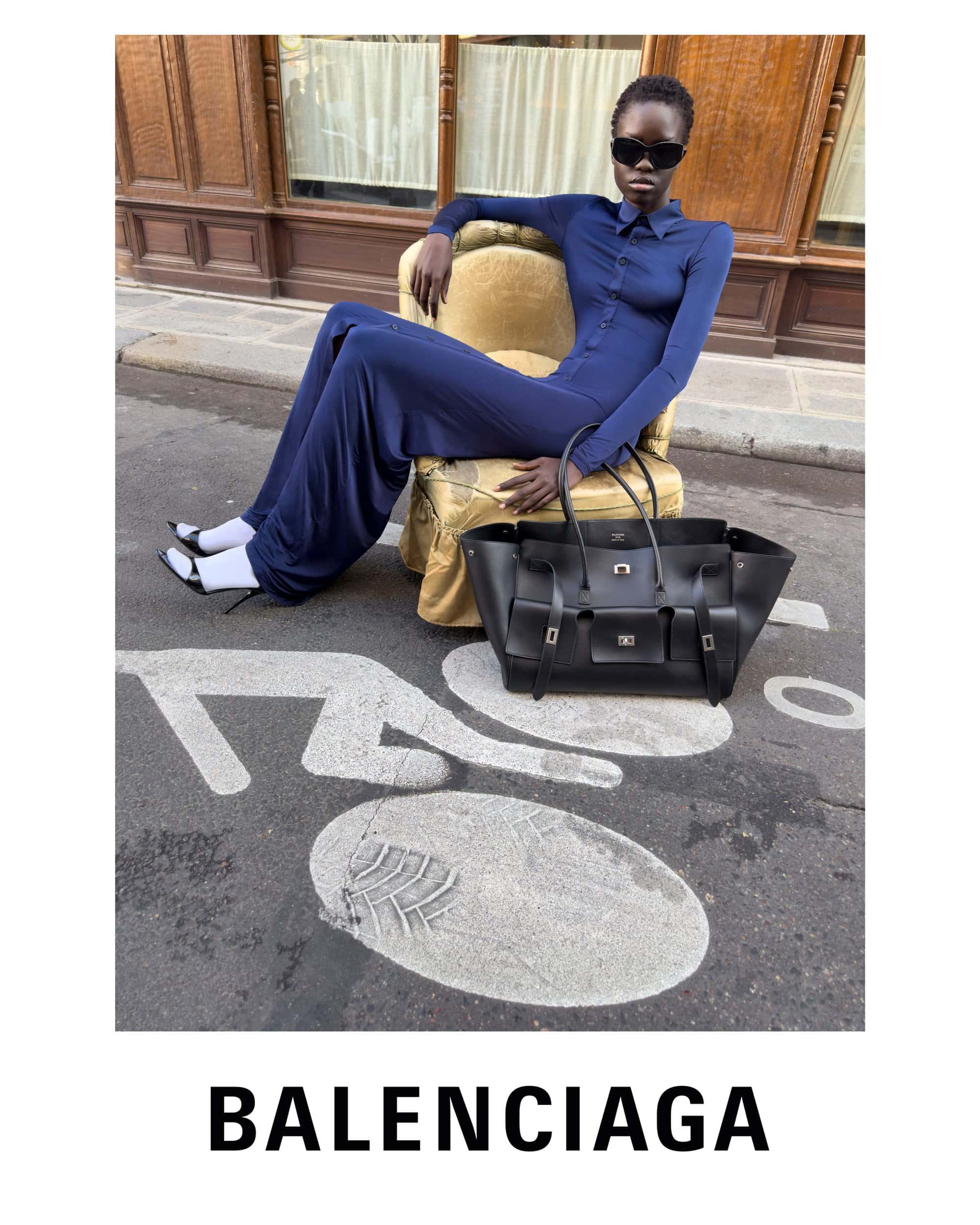 Model in blue jumpsuit wearing sunglasses seated on a gold armchair on a street next to a black leather bag for Balenciaga.