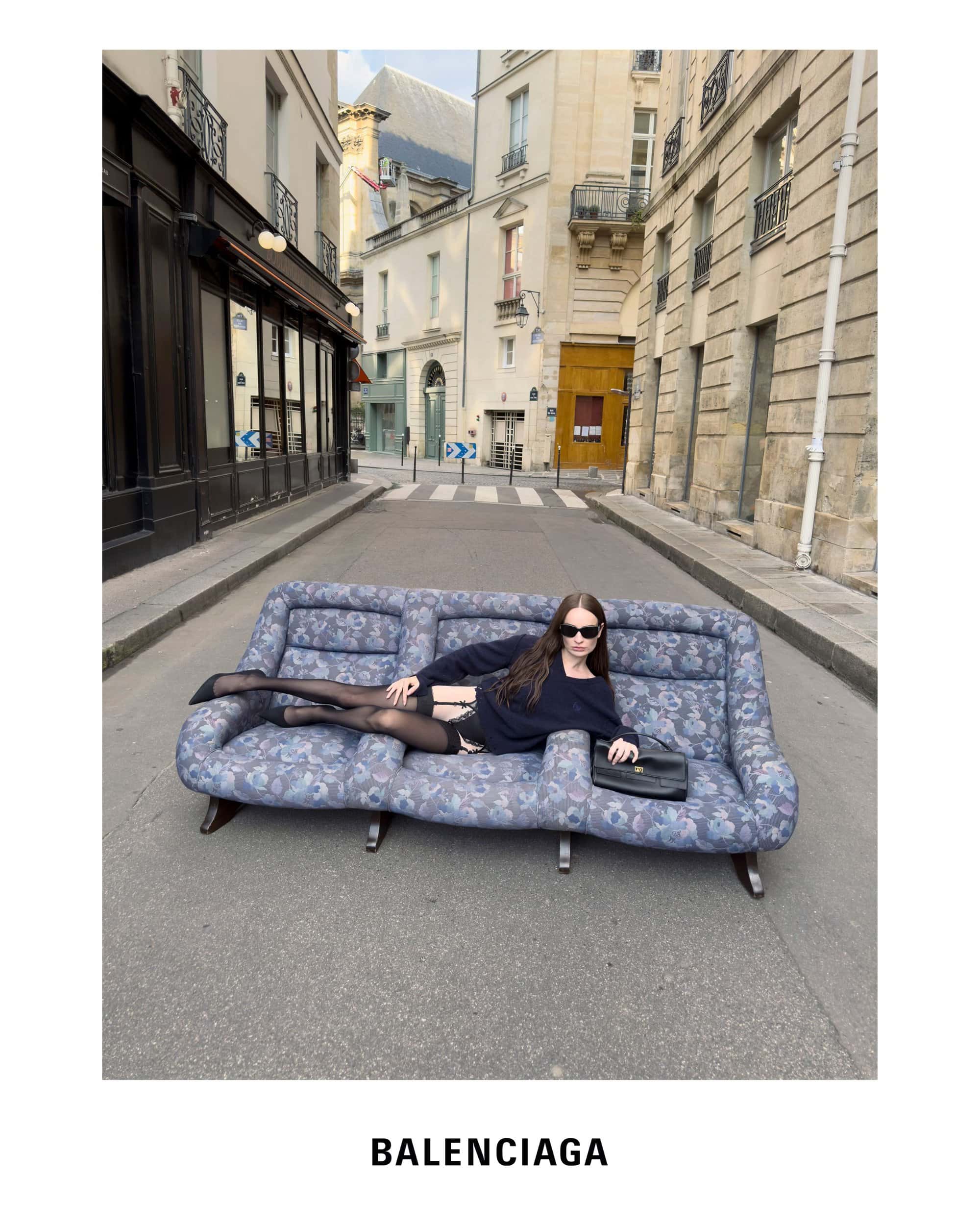 Woman wearing dark sunglasses lounging on a patterned floral sofa placed in the middle of a narrow Parisian street