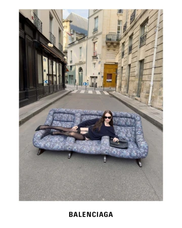 Woman wearing dark sunglasses lounging on a patterned floral sofa placed in the middle of a narrow Parisian street