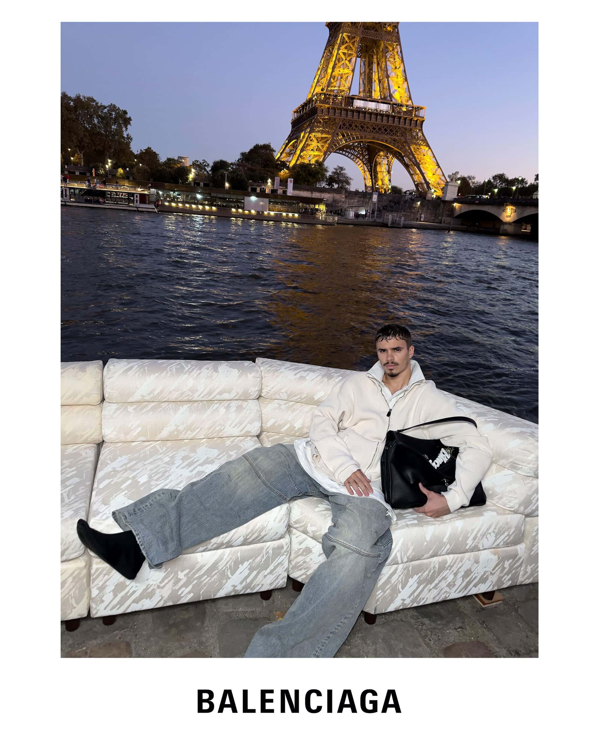 Romeo Beckham holding a black handbag seated on a white sofa outdoors with the Eiffel Tower in the background for Balenciaga