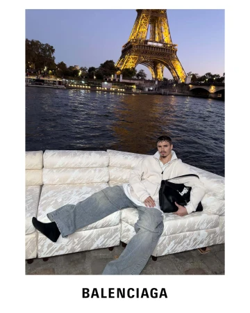 Romeo Beckham holding a black handbag seated on a white sofa outdoors with the Eiffel Tower in the background for Balenciaga