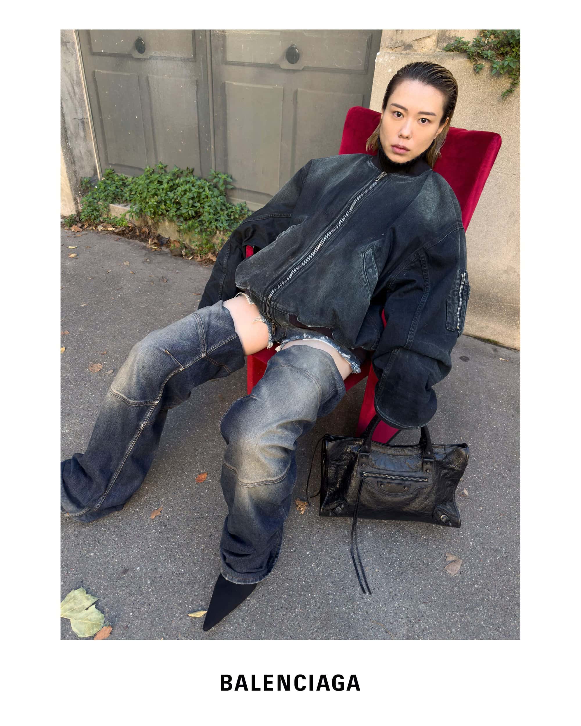 Model seated on a bright red armchair outdoors wearing an oversized dark denim bomber jacket and matching layered denim trousers.