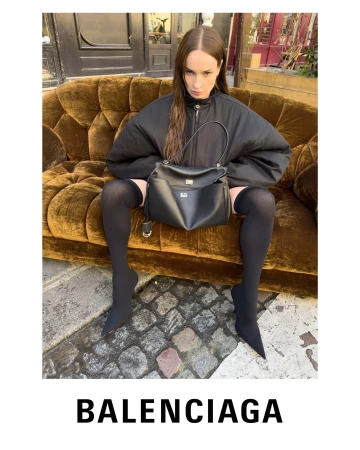 Model seated on an outdoor golden velvet sofa holding a black leather handbag against a cobblestone street backdrop