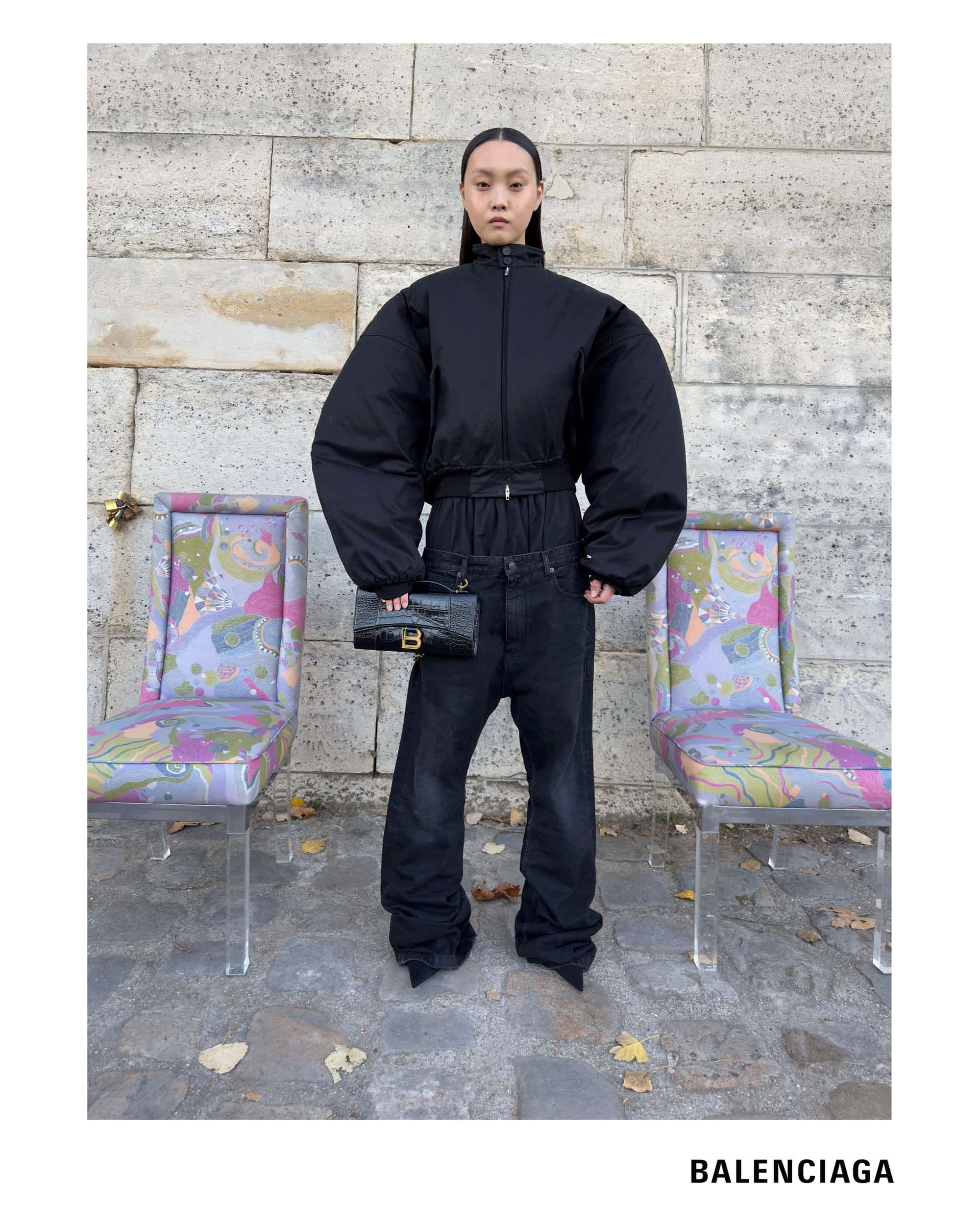 Model wearing a black oversized puffer jacket and dark jeans holding a black handbag between two patterned chairs.