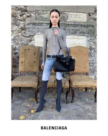 Model standing in front of stone wall wearing patterned top, jeans, and black boots while holding a large black shoulder bag.