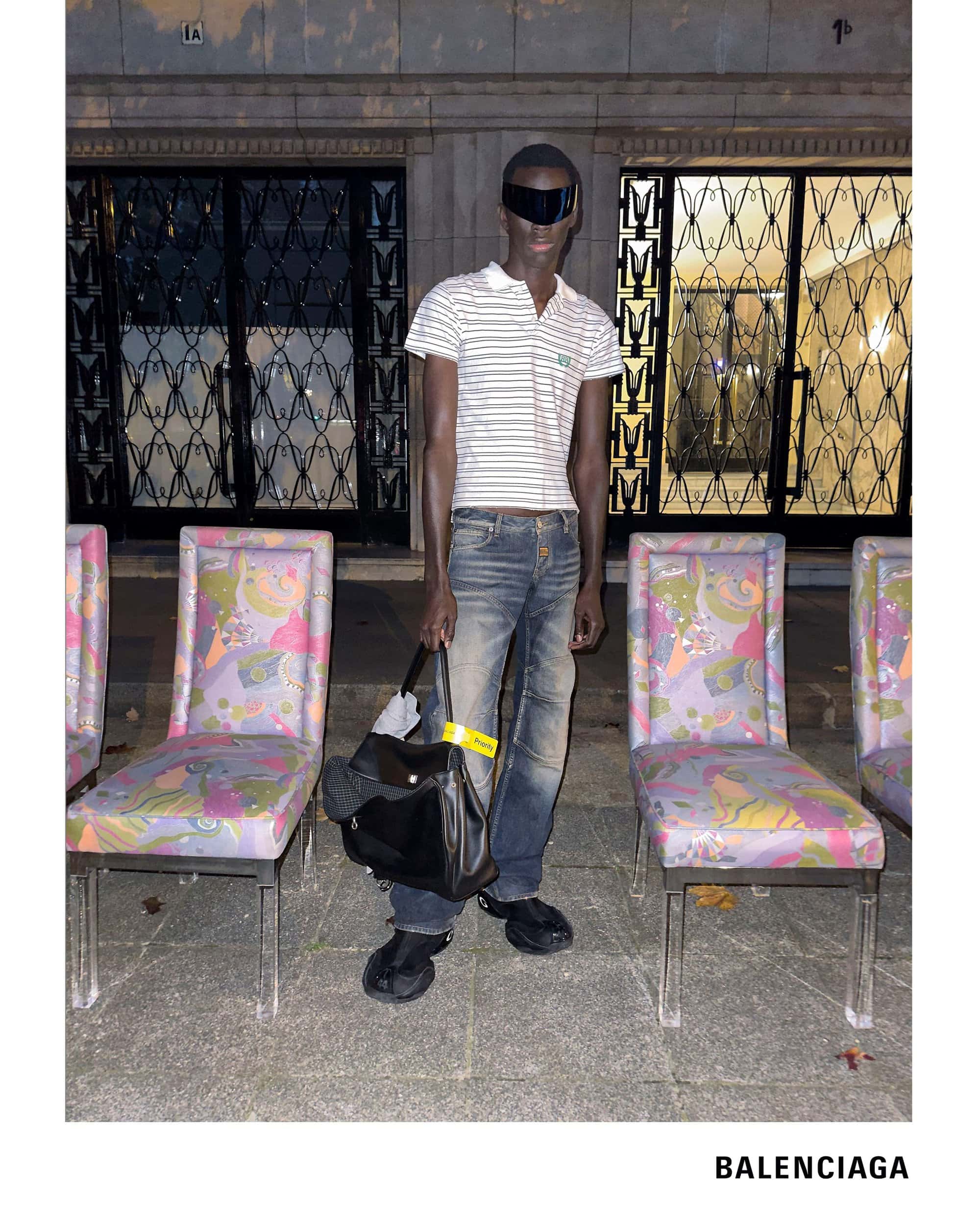 Model wearing dark sunglasses, striped polo, and distressed jeans holding a large black bag in front of patterned chairs.