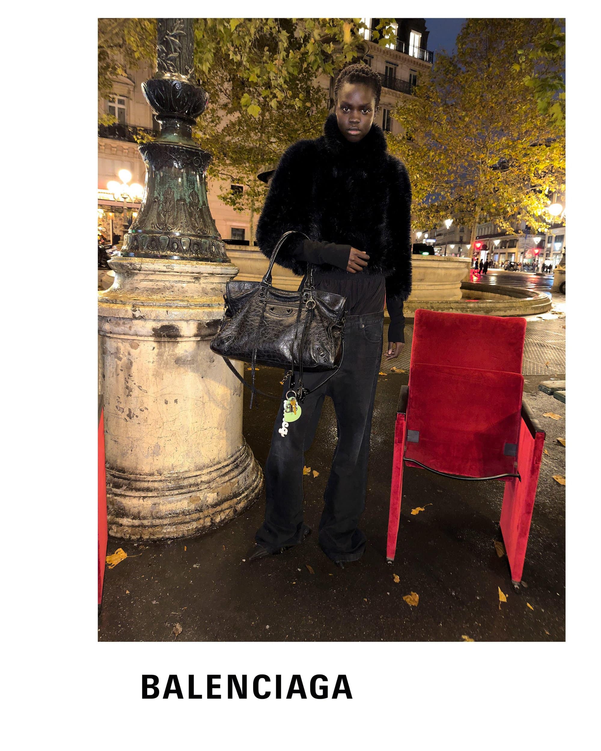 Model standing outdoors at night by a stone pillar holding a black leather handbag for Balenciaga