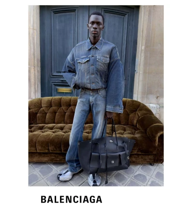 Black man wearing oversized denim jacket and jeans holding a large black leather bag in front of a brown velvet couch and dark double doors