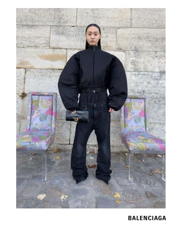Model wearing a black oversized puffer jacket and dark jeans holding a black handbag between two patterned chairs.
