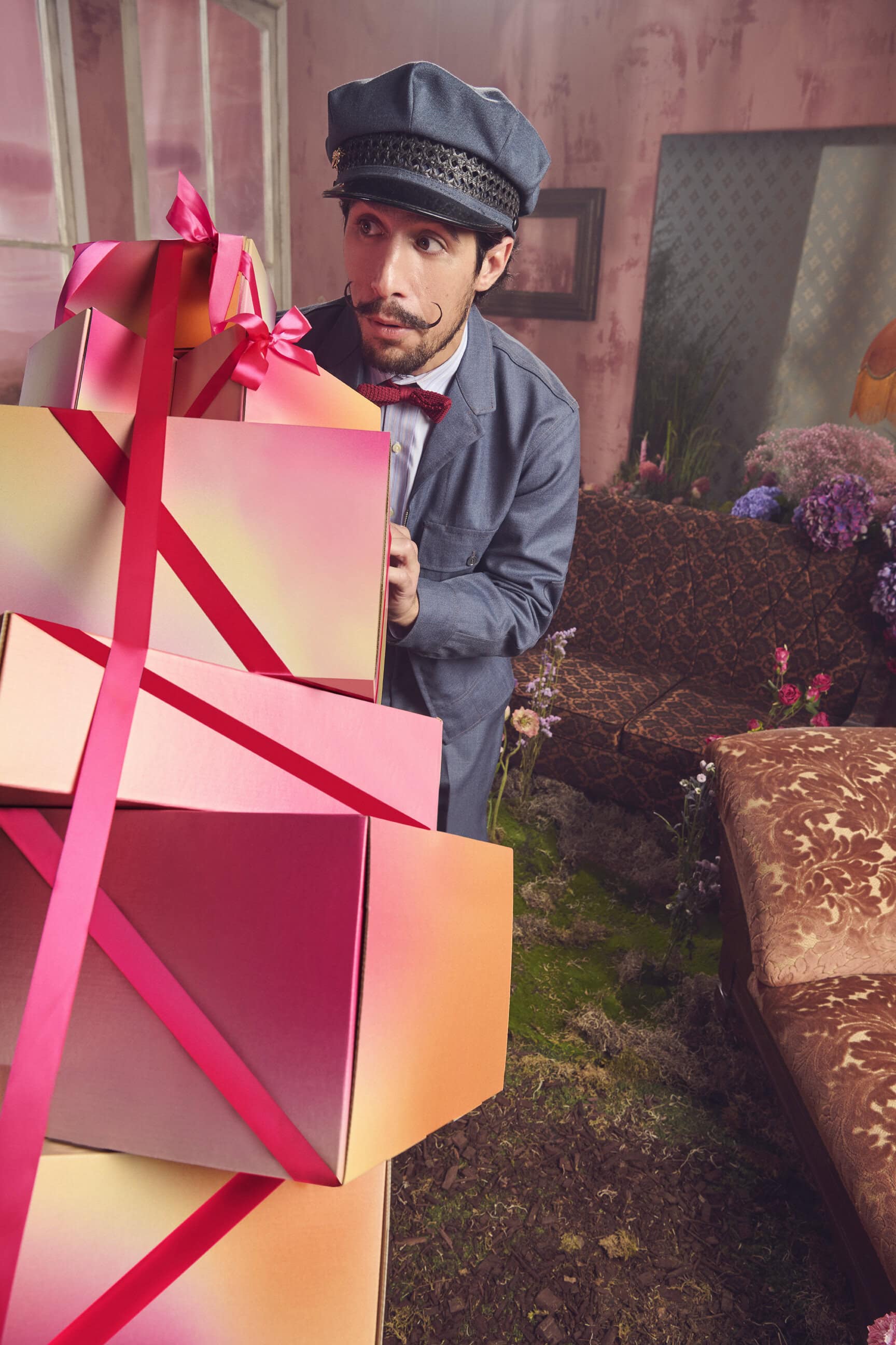 Man with mustache wearing grey suit and cap peeking behind a towering stack of gradient gift boxes tied with bright pink ribbons.