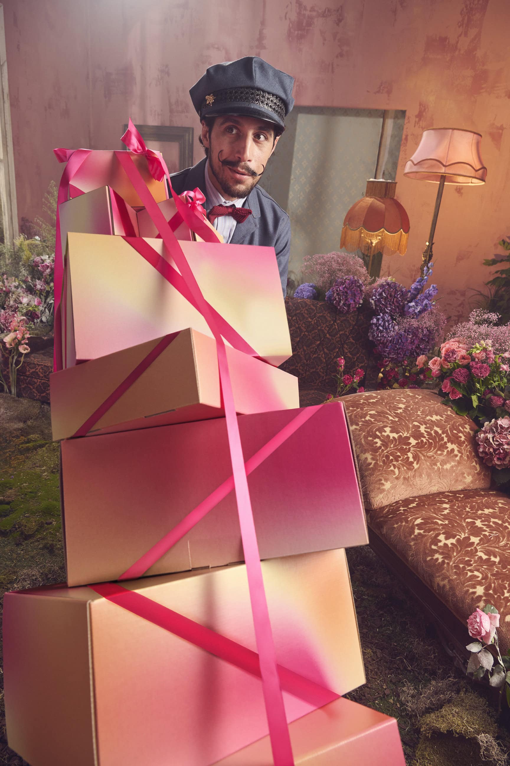 Man wearing a cap and suit standing behind a tall stack of gradient gift boxes tied with bright pink ribbons.