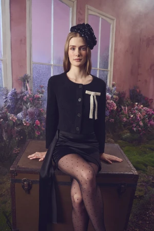 Model seated on a vintage trunk wearing a black cardigan, black skirt, patterned tights, and a dark floral hat