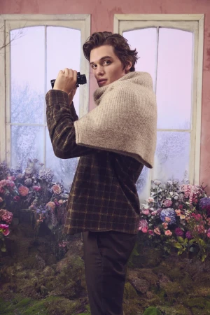 Young man holding vintage binoculars wearing plaid shirt and draped knit scarf against a floral window set