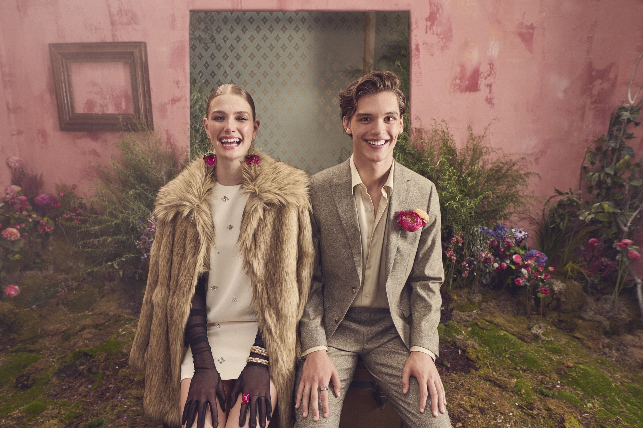 Smiling woman in a faux fur coat and a man in a tweed suit posing together against a pink textured wall with floral arrangements.