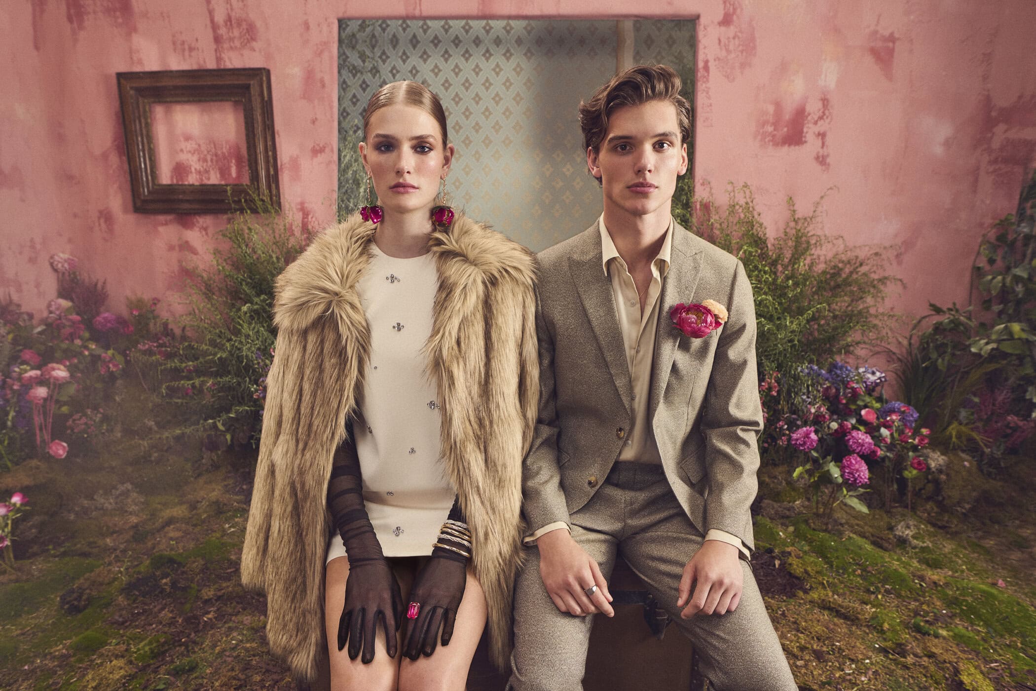 A man in a tan suit and a woman in a fur coat and white dress seated together against a pink textured wall.