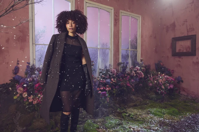Woman with curly dark hair wearing a dark coat and black dress standing before large windows inside a room filled with flowers and moss.