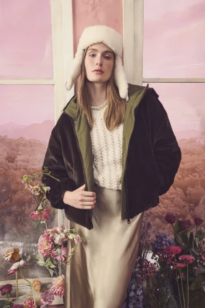 Model wearing white furry ushanka hat, dark winter coat, and satin skirt standing in front of floral window backdrop.