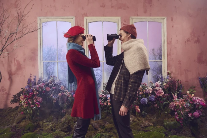 Man and woman standing indoors in front of large windows looking through binoculars amidst a floral indoor garden set