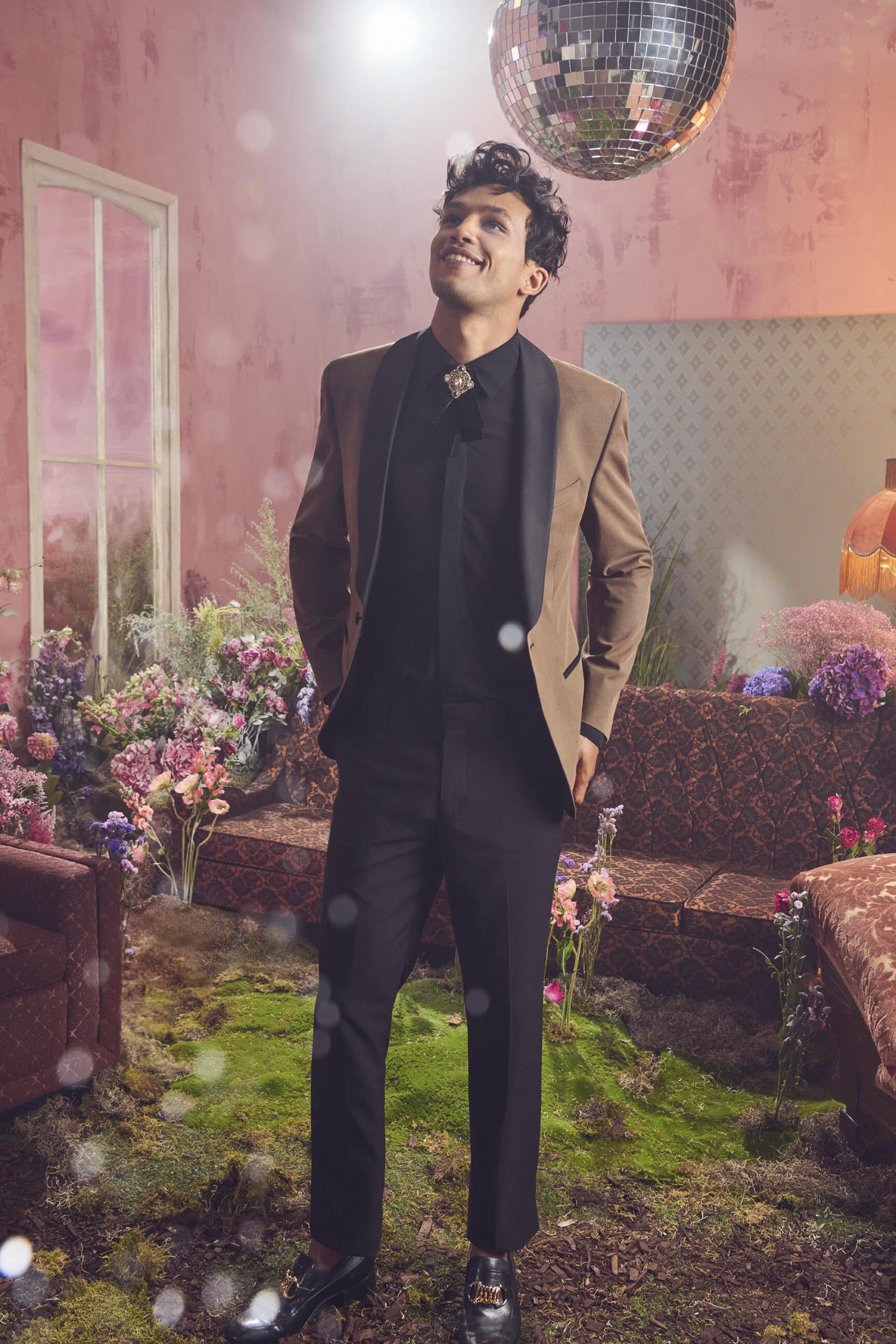 Man wearing a brown blazer and black trousers standing in an indoor set filled with flowers and a disco ball.