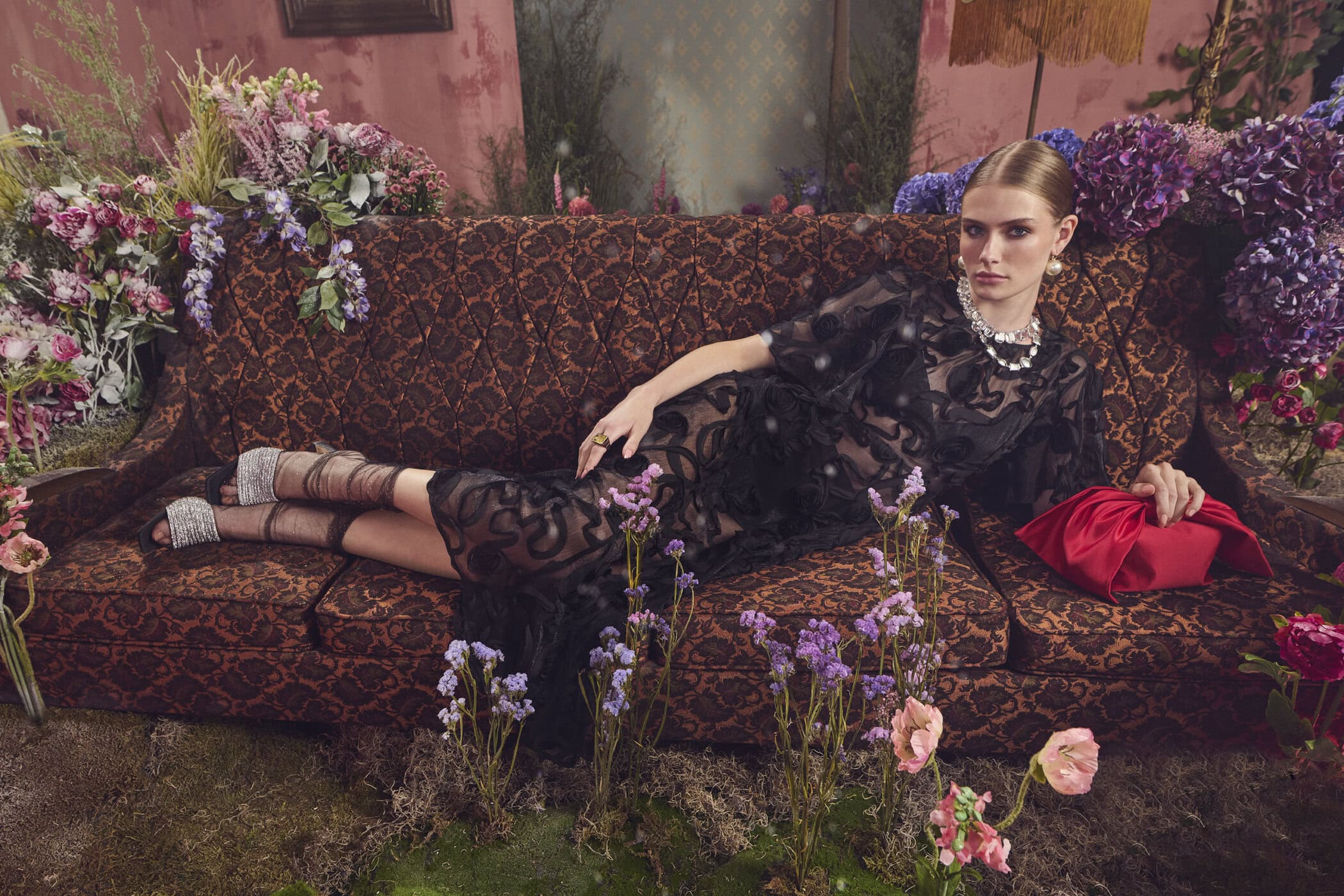 Model in black patterned dress reclining on a patterned vintage sofa surrounded by purple flowers for RW&CO.