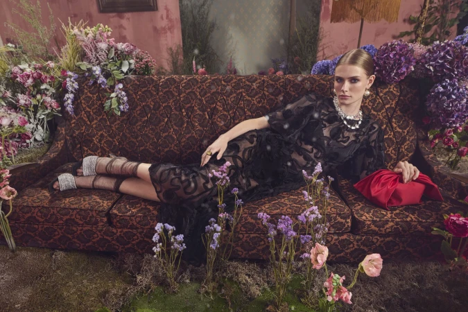 Model in black patterned dress reclining on a patterned vintage sofa surrounded by purple flowers for RW&CO.