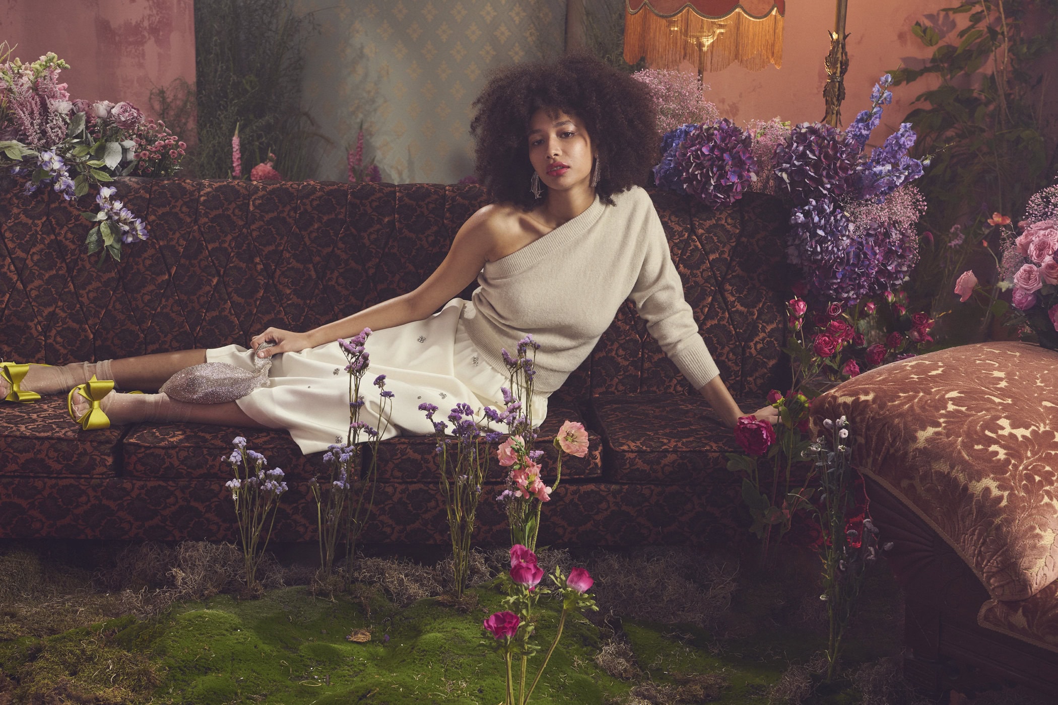 Woman with curly dark hair reclining on a patterned couch surrounded by purple flowers and mossy ground for RW&CO.
