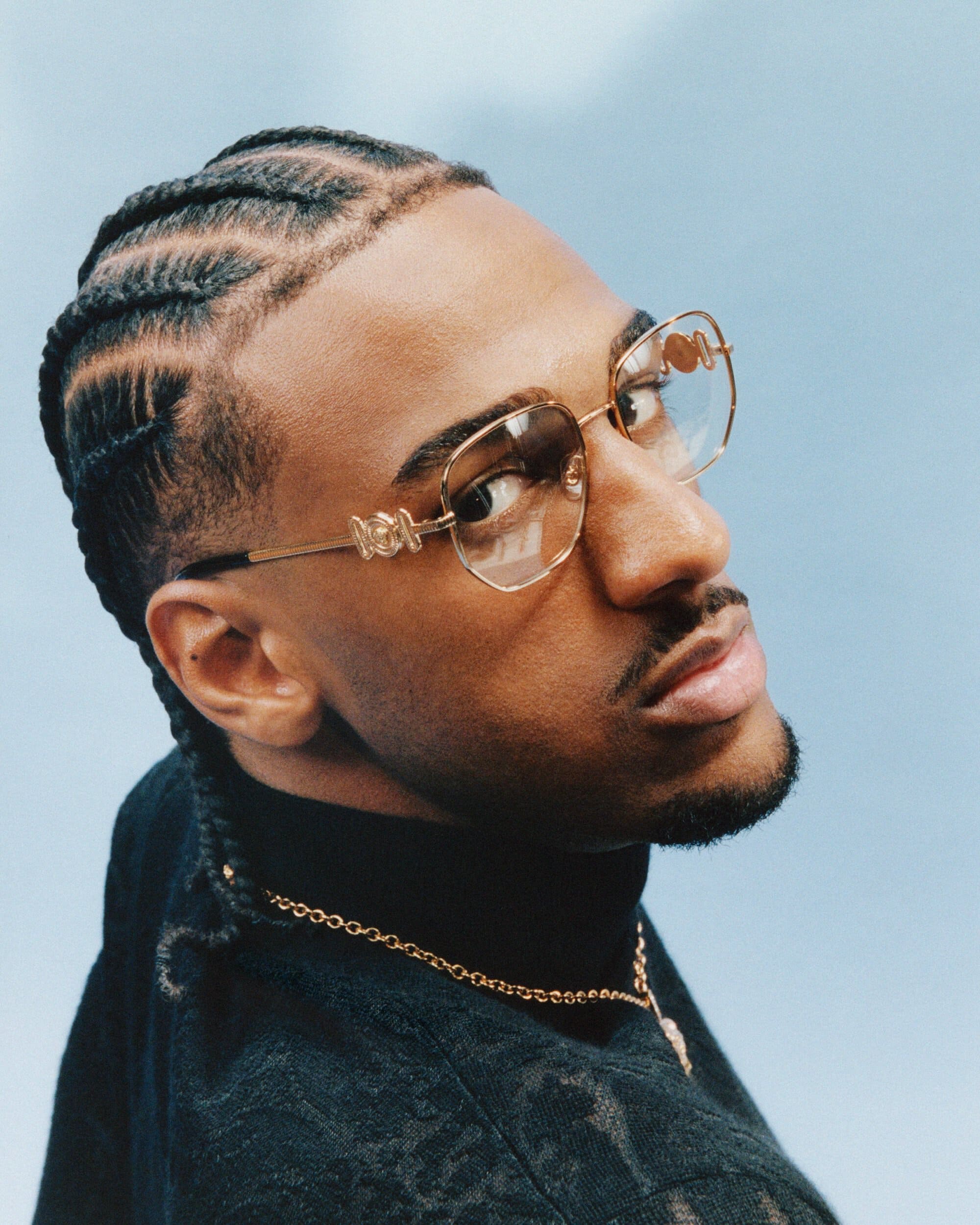 Portrait of a Black man with braided hair wearing gold-framed glasses, a black turtleneck, and a gold chain necklace against a light blue sky.