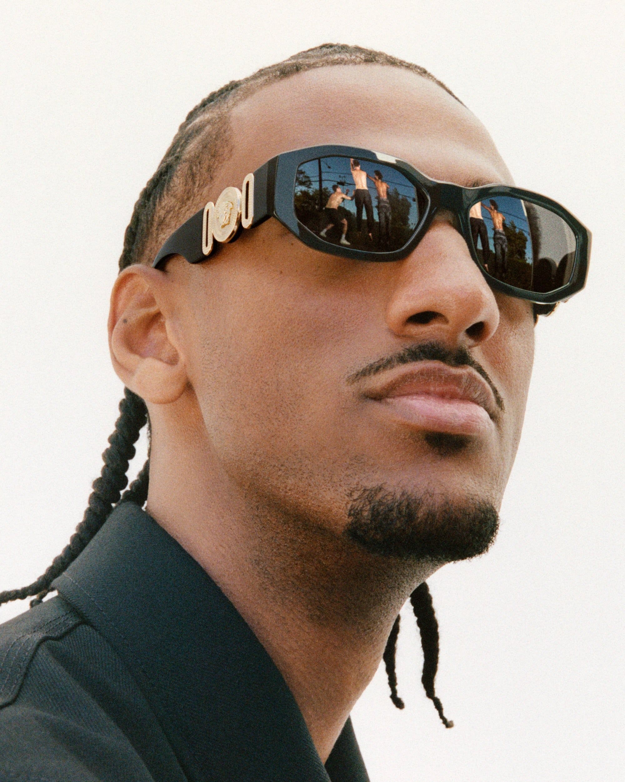 Close-up portrait of Alexandre Sarr wearing black Versace sunglasses with gold hardware against a plain background