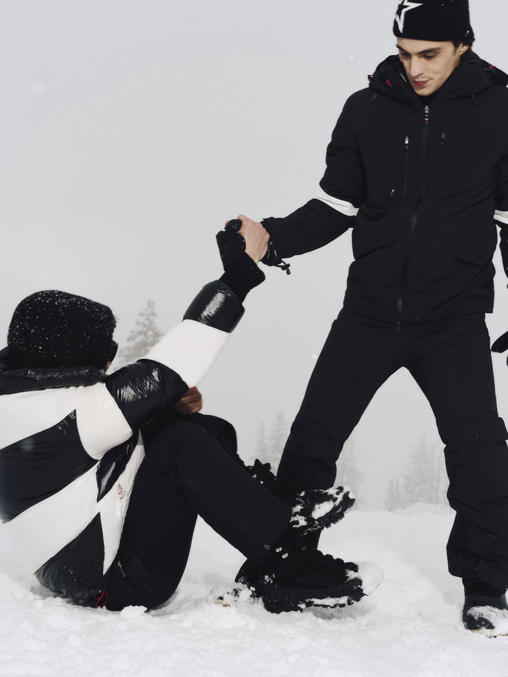 Two people in winter gear in a snowy landscape, one standing and pulling the other up from the snow.