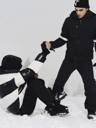 Two people in winter gear in a snowy landscape, one standing and pulling the other up from the snow.