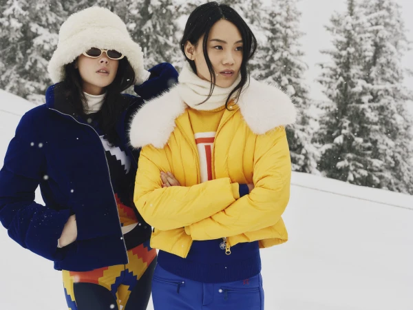 Two models wearing winter ski apparel including a yellow puffer jacket and a blue puffer jacket outdoors in snowy Gstaad, Switzerland.