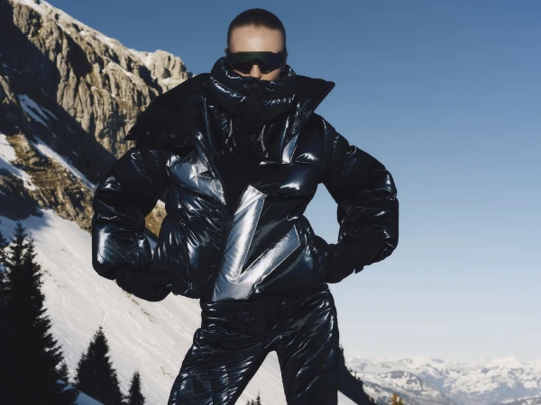 Model wearing a shiny puffer jacket and sunglasses against a snowy mountain and clear blue sky backdrop