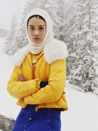 Model in a bright yellow puffer jacket with fur collar and white balaclava outdoors in a snowy landscape for Perfect Moment