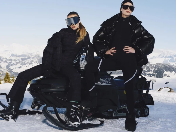 Two models in black winter apparel posing with a snowmobile against a snowy mountain landscape under a clear sky.
