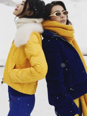 Two women standing back-to-back in snow, wearing colorful winter puffer jackets and warm scarves.
